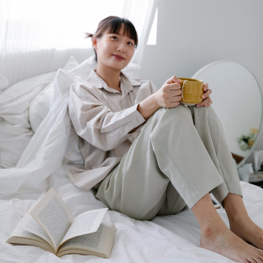 Asian American woman sitting down next to window wearing beige pajama shirt and bottom.